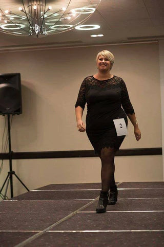 Une femme marche sur un podium en robe noire élégante, lors d'un défilé intérieur.