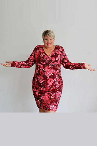 Une femme souriante pose en robe fleurie rouge, les bras écartés dans une attitude accueillante.