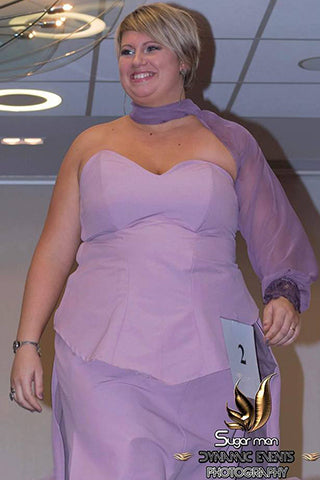 Une femme souriante défile en robe violette élégante, ornée d’un voile assorti et lumineux.