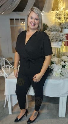 femme souriante portant une combinaison noire élégante. La combinaison a un décolleté en V, des manches courtes et une ceinture nouée à la taille, mettant en valeur sa silhouette. Elle est assortie de ballerines noires. La femme a les cheveux blonds et porte une montre dorée à son poignet gauche.